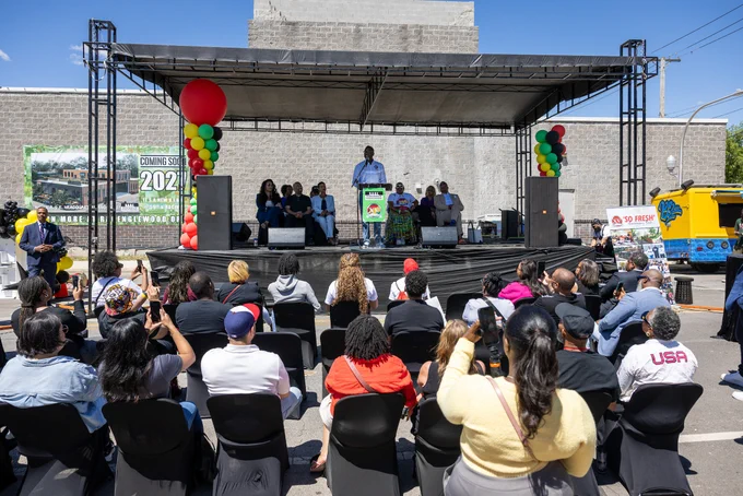 A public event with a person speaking on a stage under a canopy. The audience is seated in front, listening attentively. The stage is decorated with colorful balloons.
