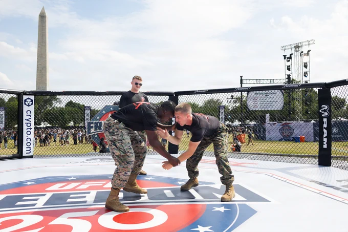 A military personnel demonstration inside a UFC-branded octagonal ring on a grassy field. The Washington Monument is visible in the background. Spectators watch the event, with various banners and equipment around the area.