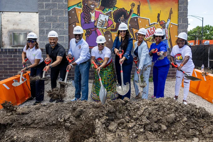 A group of people stands at a ceremonial groundbreaking event, each holding a shovel. They are wearing helmets and standing in front of a mural that depicts vibrant figures with raised fists and musical themes.