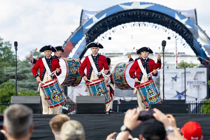 A group of musicians in historical uniforms performs on stage with drums, set against an arch decorated with stars. The scene includes an audience capturing the event, with the backdrop displaying "Freedom" and "250."