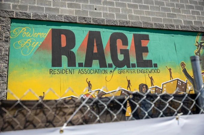 The image shows a mural on a wall with the text "Powered By R.A.G.E. Resident Association of Greater Englewood." The foreground includes a chain-link fence.