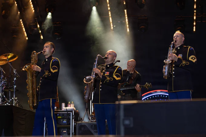 A musical band performs on stage featuring three individuals in uniform playing saxophones. Stage lights illuminate the scene, and a guitarist is visible in the background.