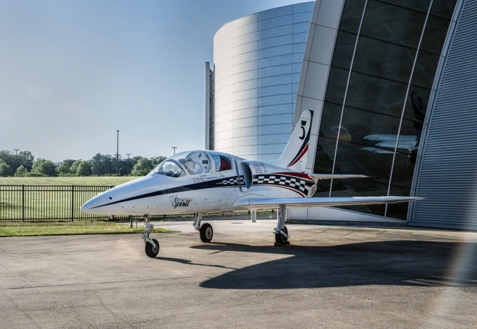 A sleek jet airplane with a polished exterior is parked in front of a modern hangar. The aircraft features a race-inspired paint design with checkered patterns.