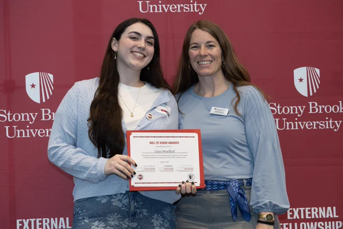 Two people stand smiling in front of a red backdrop featuring the Stony Brook University logo. One individual is holding an award certificate. Both are dressed in light blue tops.