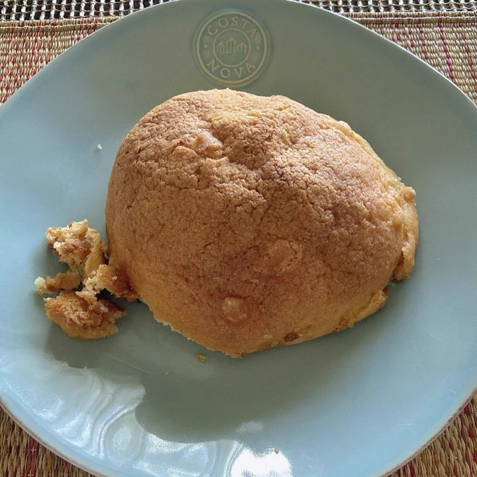 水色の皿の上に、大きなメロンパンが1つ置かれている。表面はこんがり焼き色がつき、サクサクしたクッキー生地の質感がよく分かる。左側には崩れたパン生地のかけらが少し残っており、焼きたてのような雰囲気。皿には「COSTA NOVA」のロゴが入り、編み込みのランチョンマットの上で自然光に照らされたシンプルな朝食風景を写した写真。
