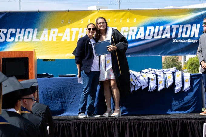 Two individuals stand on a stage at a UC San Diego Scholar-Athlete Graduation event. One person wears a graduation gown and holds a diploma. Behind them is a UC San Diego banner and a table with stoles. Audience members in graduation attire are visible in the foreground.