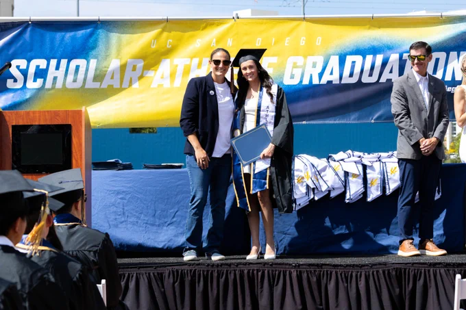 Graduates stand on stage at the UC San Diego Scholar-Athlete Graduation. One person in a cap and gown holds a diploma. A backdrop displays the event name, and people in the audience watch the ceremony.