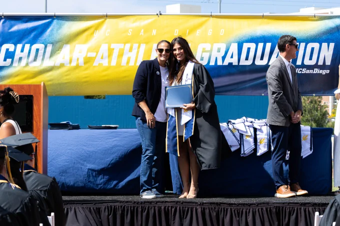 Graduates stand on stage at the UC San Diego Scholar-Athlete Graduation. One person in a cap and gown holds a diploma. A backdrop displays the event name, and people in the audience watch the ceremony.