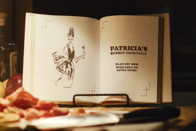 From Widow’s Bay. A book is open on a stand with a knife and food in front of it. On the left page of the book is a drawing of a woman holding a glass in her left hand. Text on the book reads “Patricia’s Sunset Cocktails.” Below that text reads “Playlist now available on Apple Music.”