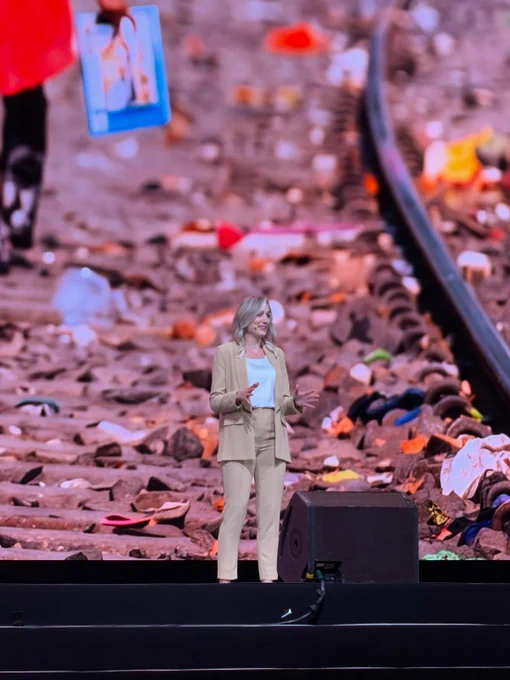 A speaker stands on stage at the Rotary International Convention in Taipei, presenting in front of a large screen displaying a close-up photograph of railroad tracks with scattered debris. The speaker gestures while addressing the audience.