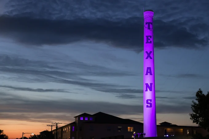The image shows a tall, cylindrical tower illuminated in purple light. The word "TEXANS" is vertically displayed on the tower. Buildings and trees are visible at the base, with a darkening sky and a sunset on the horizon.