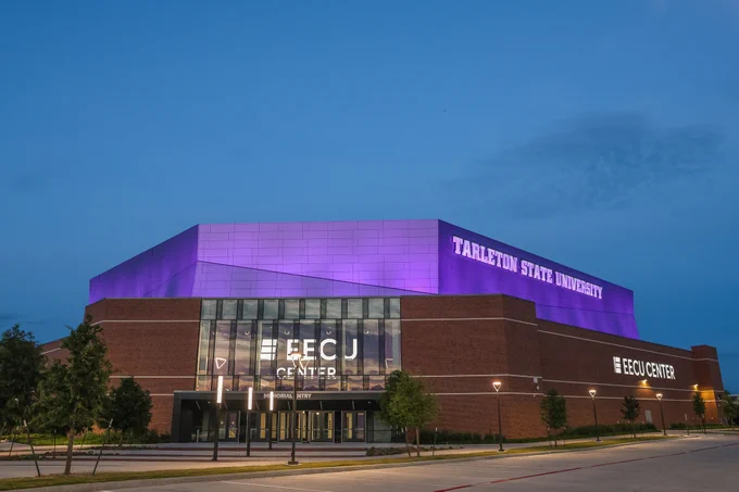 The image shows the EECU Center at Tarleton State University. The building is illuminated with bright purple lighting on its upper facade. The sky is clear, and the area around the building is well-lit.