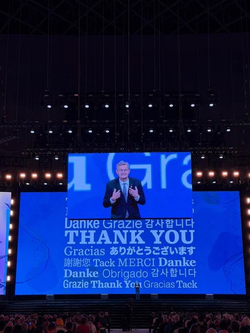 Holger Knaack speaks on stage during a Rotary International Convention general session. A large screen behind him displays “Thank You” in multiple languages as attendees watch from the audience.