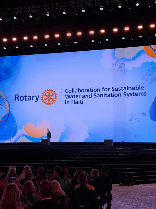 Wide view of the main stage during a Rotary International Convention general session in Taipei. A large screen displays the newly announced sixth Programs of Scale grant, “Collaboration for Sustainable Water and Sanitation Systems in Haiti,” alongside the Rotary logo. Rotary Foundation Chair Holger Knaack stands on stage as attendees watch from the audience.