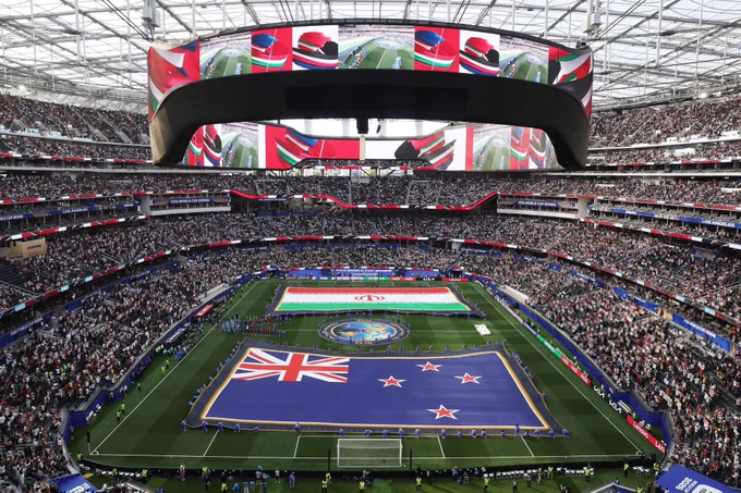 لقطة نهارية واسعة بزاوية مرتفعة تستعرض الأجواء الاستثنائية داخل استاد "صوفي" (SoFi Stadium) في لوس أنجلوس، أثناء مراسم ما قبل انطلاق مباراة في بطولة كأس العالم 2026 تجمع بين منتخبي نيوزيلندا وإيران. يمتد على العشب الأخضر للميدان علمان قماشيان عملاقان؛ علم نيوزيلندا الأزرق الذي يضم علم الاتحاد البريطاني والنجوم الحمراء الأربعة في مقدمة الأسفل، وعلم إيران بألوانه الأخضر والأبيض والأحمر وشعاره الوطني في الأعلى، ويتوسطهما شعار البطولة الدائري. يبرز المعمار الداخلي الفاخر للاستاد المزدحم بعشرات الآلاف من المشجعين في المدرجات متعددة الطوابق، وتلفت الأنظار الشاشة الإلكترونية الدائرية المعلقة الضخمة والأيقونية (Infinity Screen) التي تطوق سقف الاستاد بالكامل، وهي تعرض لقطات ملونة مكررة للأعلام لإضفاء طابع بصري حماسي ومبهر على حفل التدشين.