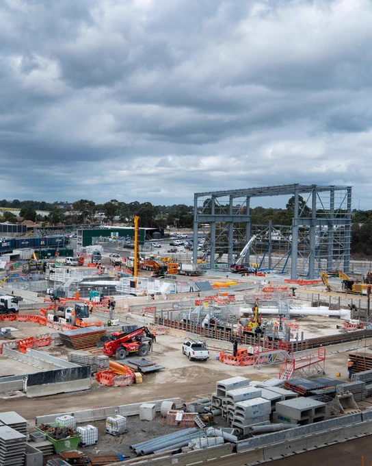 A construction site with various machinery and equipment, including cranes and scaffolding. There are numerous building materials and protective barriers scattered throughout the area. The sky is overcast with thick clouds.