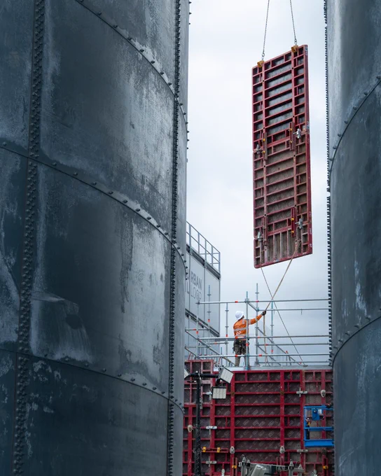 A person works on scaffolding at a construction site, surrounded by large metal structures. A red metal framework is being hoisted by a crane. The scene conveys a sense of industrial activity.