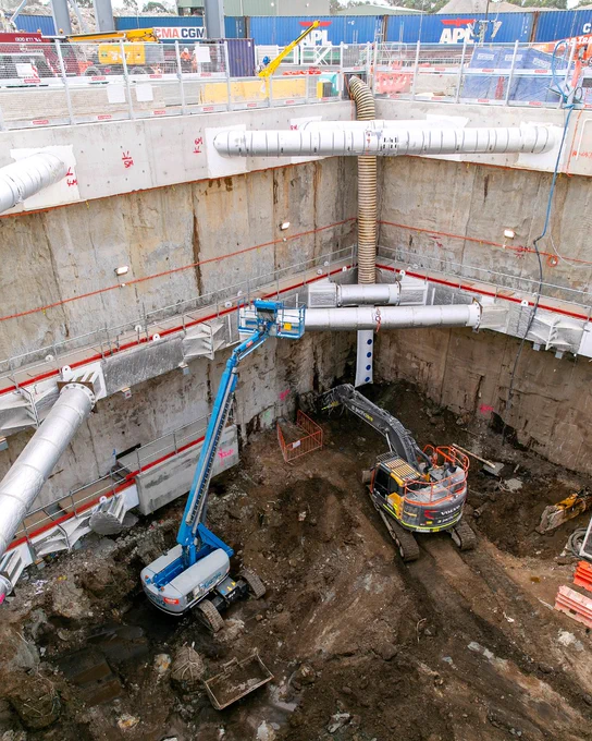 A construction site with a deep excavation area. Two construction machines are present: a blue articulated lift and a yellow excavator. The area is surrounded by large, curved pipes and concrete walls. Safety barriers and equipment are visible, along with some shipping containers in the background.