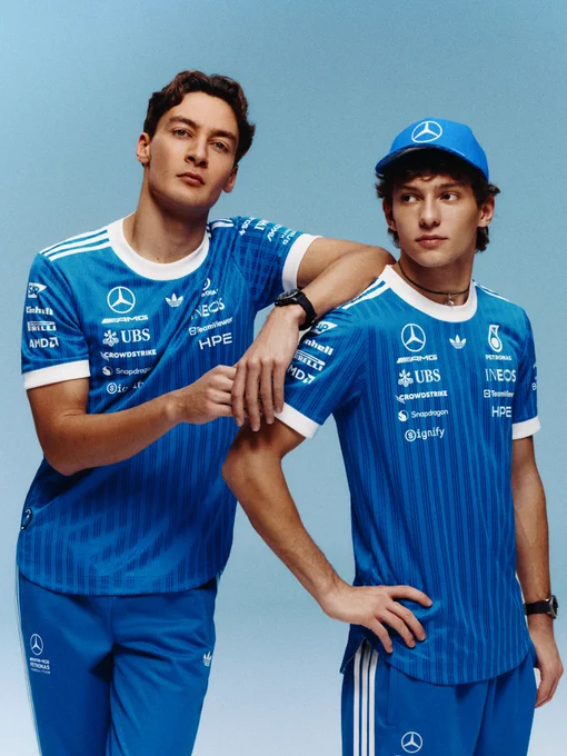 George Russell and Kimi Antonelli stood in front of a light blue backdrop. Both wearing blue thinly striped Mercedes Blue Wonder shirts and matching blue tracksuit bottoms. Kimi is stood with one hand on his hip, looking out to his right. George is leaning on Kimi's shoulder beside him.