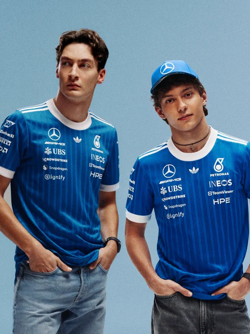 George Russell and Kimi Antonelli stood in front of a light blue backdrop. Both wearing blue thinly striped Mercedes Blue Wonder shirts and jeans. Both have their hands in their pockets. Kimi is looking towards the camera and George off to his right.
