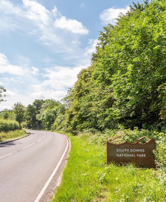 South Downs National Park sign on side of the road with greenery and a cloudy blue sky.