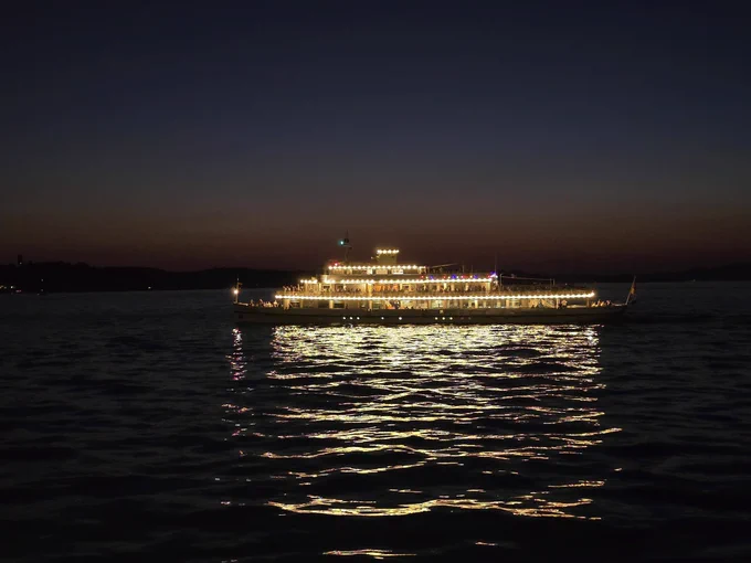Zu sehen ist ein hell erleuchtetes Ausflugsschiff. Mehrere Lichterreihen sorgen dafür, dass es hell ist. Die Lichter spiegeln sich im Wasser. Im Hintergrund ist ein letzter Rest der Abenddämmerung zu sehen, bevor es ganz dunkel wird. 
