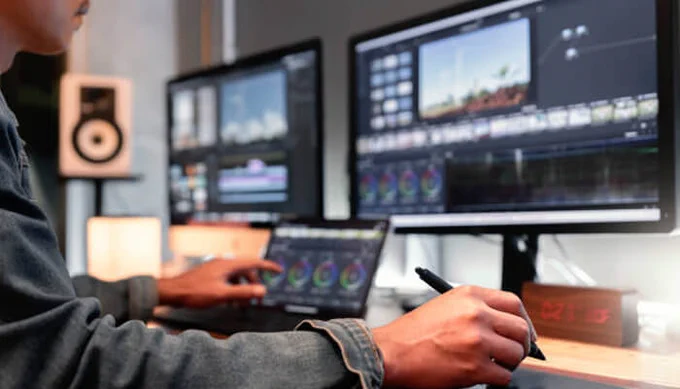 Person working on video editing and color grading on dual monitors, using a pen tablet at a modern editing workstation, focused on professional digital content creation.