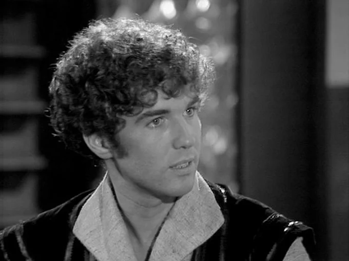 GPT 5.5 describes the image as: "A black-and-white production still from the classic BBC television series Blake’s 7, showing actor Steven Pacey in character. He is framed in a close-up, turned slightly to one side with an alert, intent expression, as if reacting to something or someone just off-camera. His curly hair, youthful features, and focused gaze stand out strongly in the high-contrast lighting. He wears a futuristic-looking costume with a broad, pale collar beneath a darker outer garment, fitting the series’ distinctive low-budget but imaginative science-fiction style. The background is softly blurred, suggesting an interior spaceship or base setting, with shadowy structures and small highlights that give the scene a dramatic, studio-lit atmosphere."
The script search url for this tweet is
https://blake.torpidity.net/s/306/116