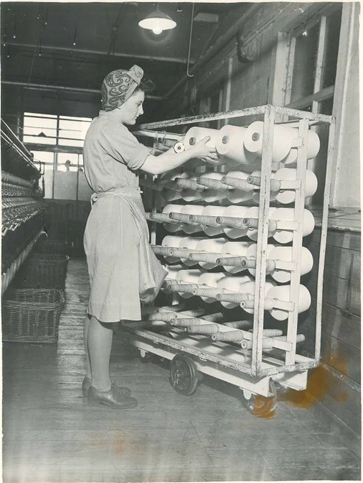 Woman in a factory, wearing a headscarf, places a spool of thread onto a trolley.