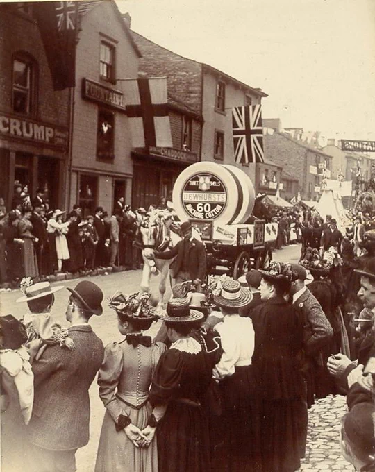 A parade float with a giant spool of Dewhurst's cotton, pulled by a horse, with crowds watching on a street decorated with flags.