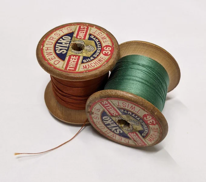 Two vintage wooden spools of sewing thread, one rust-coloured and one green, with labels reading "DEWHURST'S SYLKO".