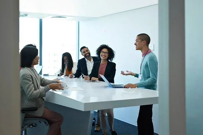 A group of Black and Asian employees in professional work clothing standing around a white desk with a laptop.