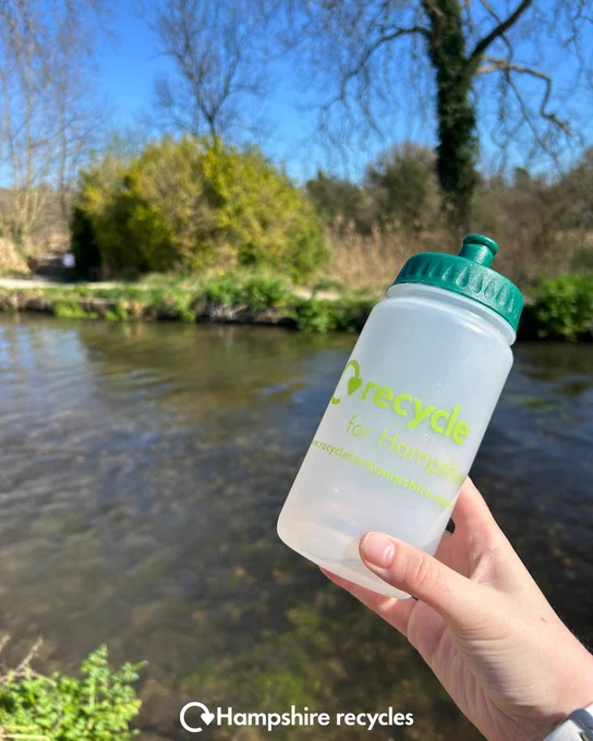 Alt text: Hand holding a clear water bottle with a green lid and Hampshire Recycles logo in front of a river; white Hampshire Recycles logo at bottom centre.