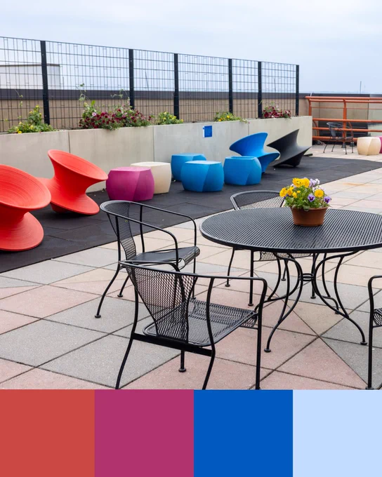 A rooftop patio features modern, colorful furniture. Red, pink, blue, and black spinning chairs line one side, contrasting with traditional black metal chairs and tables. A round table with a potted flower centerpiece sits prominently in the foreground. A low concrete wall with flower planters and a grid metal fence frames the background. The patio floor is made of large, square tiles in neutral tones, and a cloudy sky is visible above.