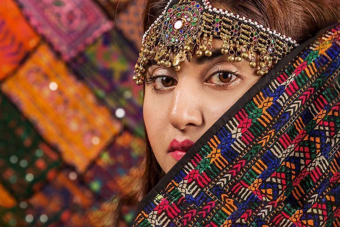 An Indian woman in colorful traditional dress.  She has large brown eyes, a scarf and a head decoration.