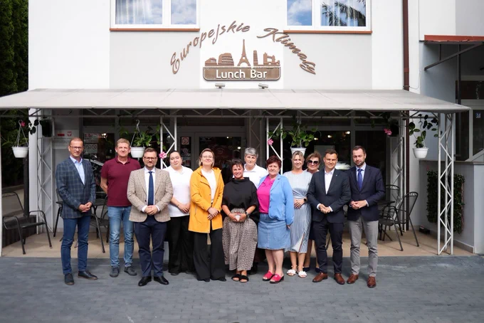 Spotkanie z lokalnymi NGO z podkarpacia. Szeroki kadr, zdjęcie grupowe. Grupa dwunastu osób pozuje do wspólnego zdjęcia przed wejściem do restauracji „Europejskie Klimaty” w Jarosławiu. W centrum kadru widoczna Sekretarz Stanu Adriana Porowska. 