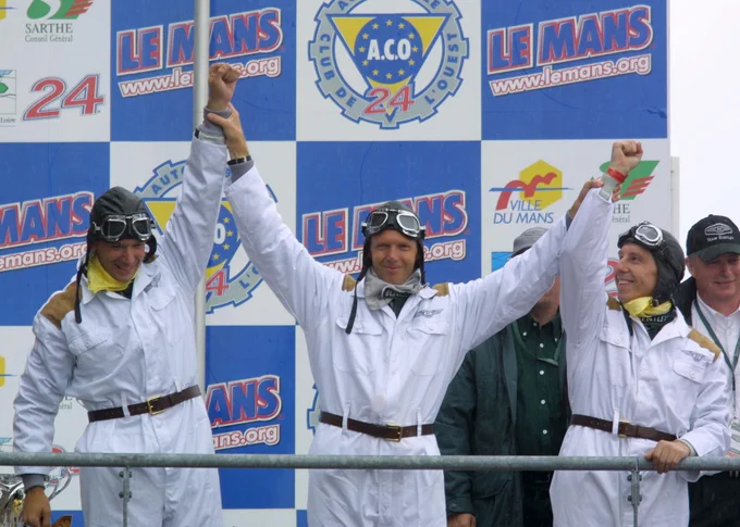Butch Leitzinger, Eric van de Poele and Andy Wallace stand on the podium; van de Poele in the centre holds up the arms of his co-drivers, all three wearing race outfits harkening back to their 1930 win: plain white overalls, brown belts, leather racing caps, goggles positioned on their foreheads and neckerchiefs. 2001 Le Mans 24 Hours, Clive Rose/LAT Images