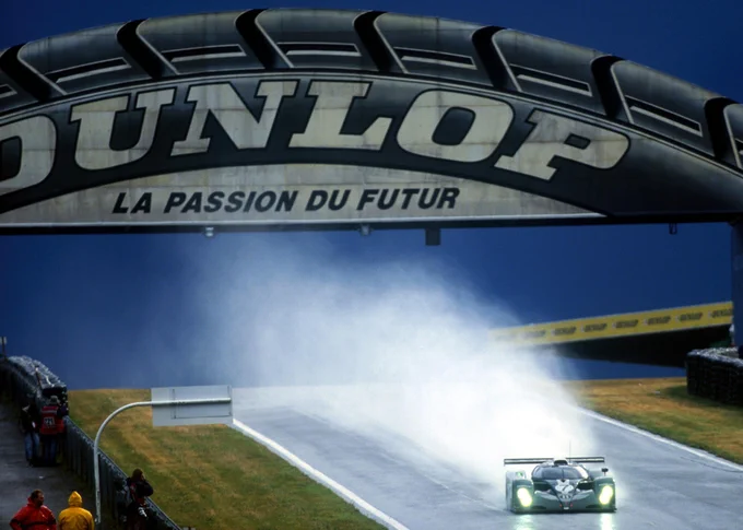 Small in the bottom right of frame, the green Bentley EXP Speed 8 passes beneath the large Dunlop Bridge, a cloud of white spray filling the air behind. 2001 Le Mans 24 Hours, James Bearne/Sutton Images