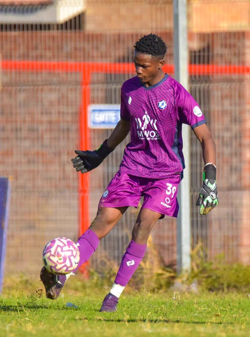 A soccer player wearing a purple jersey and gloves kicks a ball during a game. The player appears focused on the action.
