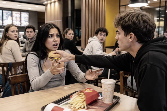 Eine Frau schaut entsetzt auf den Mann ihr gegenüber, der ihr den Burger aus der Hand nimmt. Andere Gäste im Restaurant beobachten diese Szene.