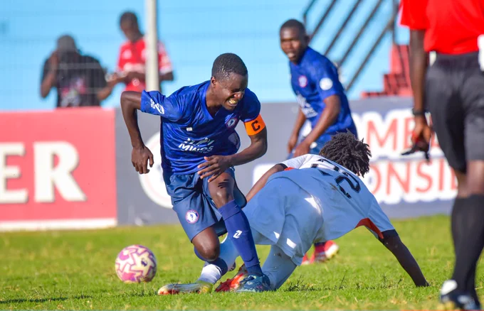 A tense moment during a soccer match with a player in blue struggling to maintain control of the ball while another player in white is on the ground. The atmosphere is competitive and dynamic.