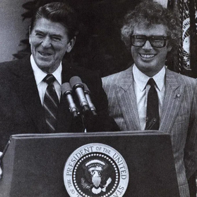 Ronald Reagan stands at a podium with the presidential seal and microphones in front of him, smiling during an outdoor event. Ken Taylor stands beside him, also smiling, wearing glasses and a light pinstriped suit. An American flag is partly visible behind them.
