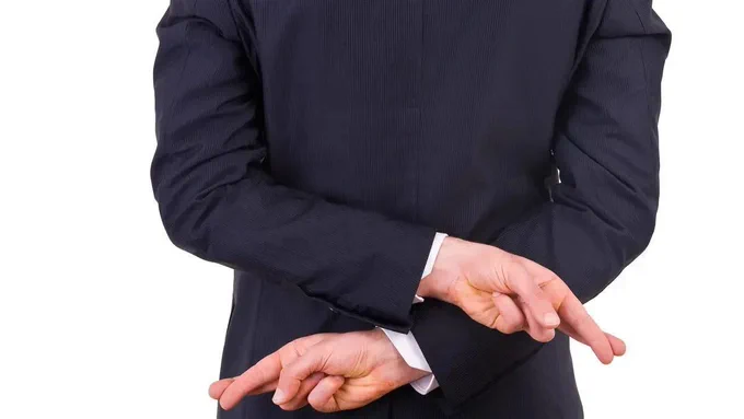 A white man in a navy blue suit, with his hands and finggers crossed behind him denoting deception.