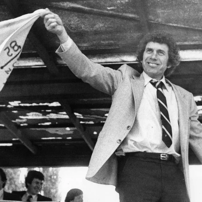 Roger Neilson smiles while standing under a stadium roof, wearing a light suit jacket, dress shirt, striped tie, and dark trousers. He raises one arm high and holds a small cloth or towel with markings on it. Several people in suits are seated behind him in the background.
