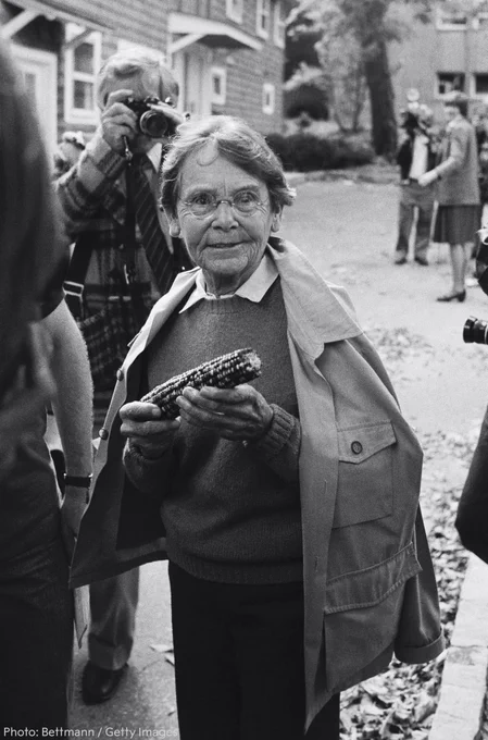 Barbara McClintock holding an ear of corn with a smile, as other individuals, including one with a camera, look on in the background.