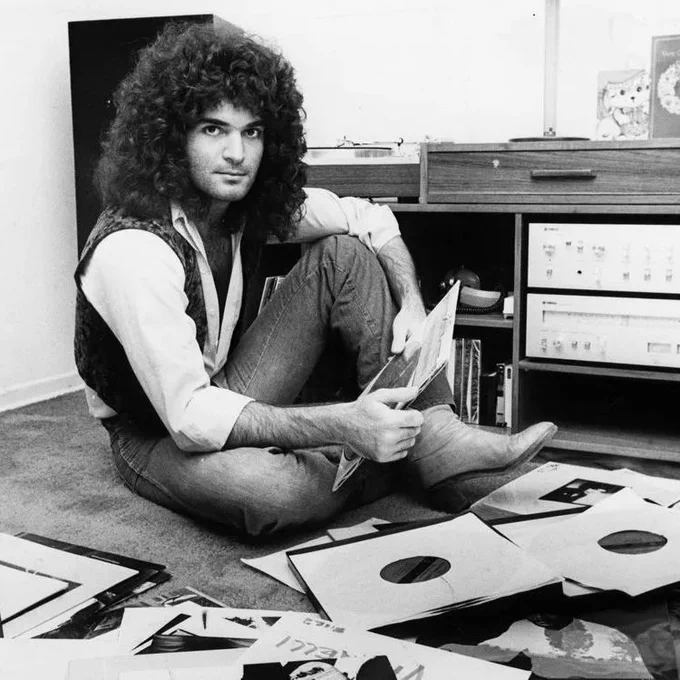 Gino Vannelli sits cross-legged on a carpeted floor, holding a record sleeve while surrounded by scattered vinyl records and album covers. He has long curly hair and wears a light shirt, vest, jeans, and boots. A stereo cabinet, shelves, books, and audio equipment are visible behind him.
