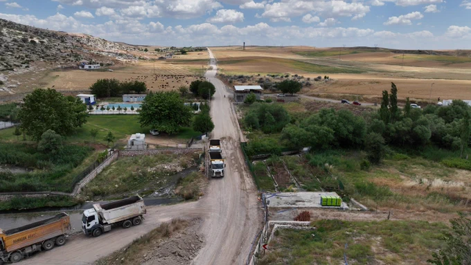 Yüksekten çekilmiş görüntüde, ortadan geçen açık renkli bir yol kırsal alanda ufka doğru uzanıyor. Yol üzerinde ve alt bölümde birkaç kamyon bulunuyor. Yolun iki yanında tarım arazileri, ağaçlık alanlar, küçük yapılar ve araçlar yer alıyor. Sol tarafta kayalık yamaç görülüyor. Gökyüzü geniş bir alanı kaplıyor ve dağınık bulutlarla örtülü.