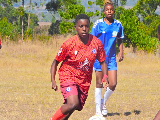 Two young soccer players compete on a grass field. One in red is dribbling the ball while the other in blue follows closely. The scene is energetic and vibrant.