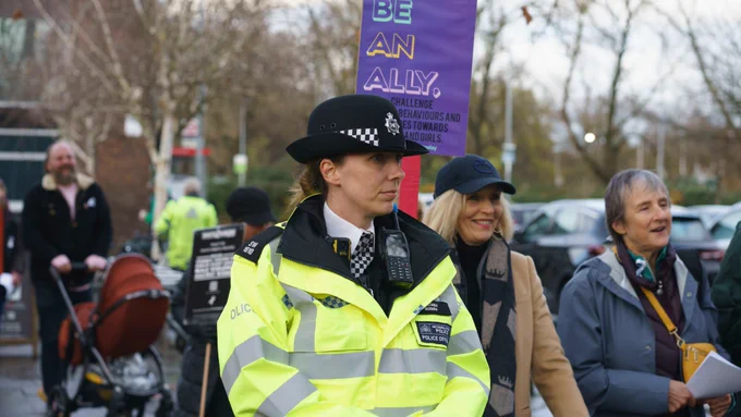 A police officer in a high-visibility jacket and hat stands at an event with a banner promoting allyship.