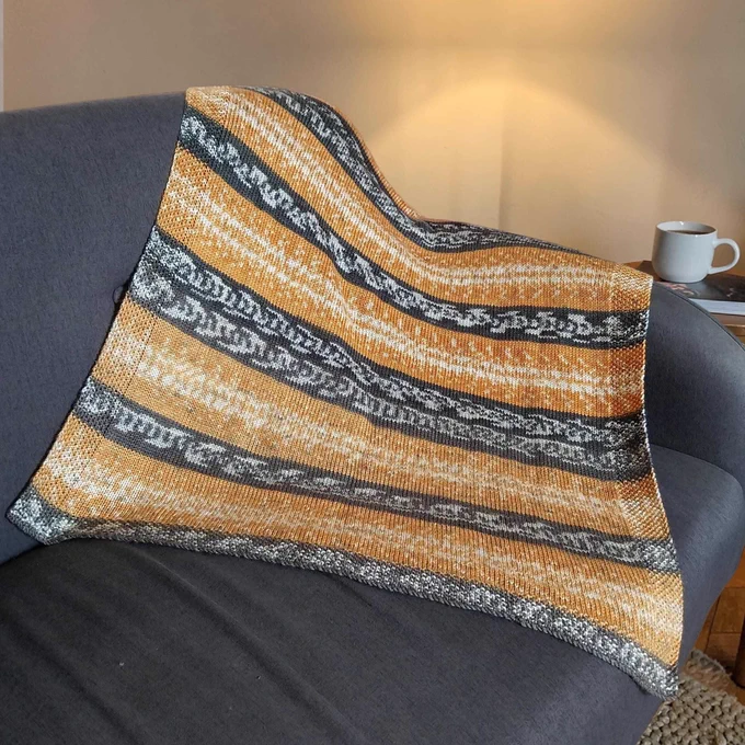 Knitted blanket with golden yellow and dark grey patterned stripes draped on a dark sofa. A warm light glows on the wall behind. To the right, a white mug sits on a wooden side table. The scene is cosy and minimalist.